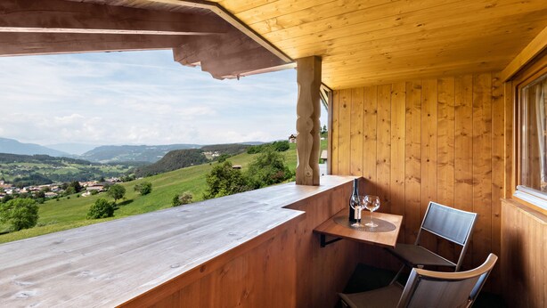 A wooden balcony with a table and chairs, overlooking a green landscape with distant houses.