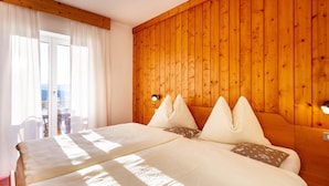 Cama doble con ropa de cama blanca, cabecera de madera y vistas al exterior a través de una ventana con cortinas blancas.