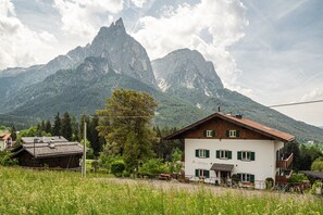 A mountain landscape with a white building featuring green shutters, a wooden roof, and a balcony.