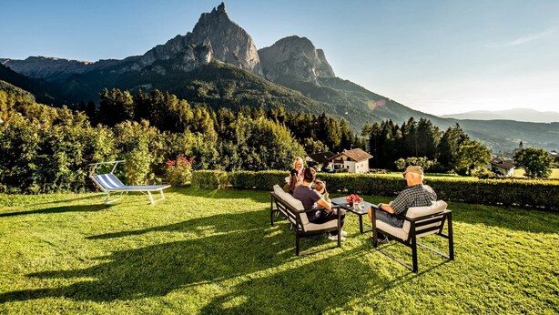 Un grupo de personas sentadas en muebles de exterior en una zona cubierta de césped, con vistas a una montaña.