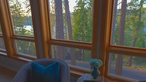 Ein Zimmer mit einem großen Fenster, das einen Blick auf hohe Bäume und ein Gewässer freigibt. Ein Stuhl mit blauem Kissen und ein kleiner Tisch mit einer Vase und Büchern sind vorhanden.