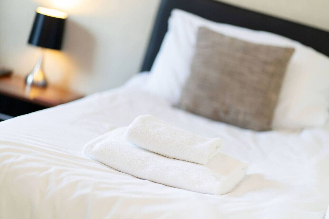 A neatly made bed with a folded white towel, a white pillow, and a dark headboard.