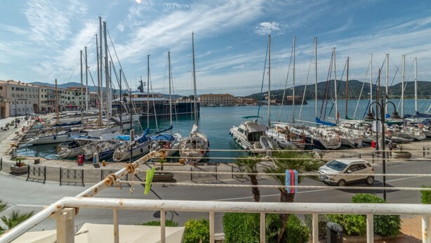 Un puerto deportivo con barcos amarrados, una carretera en primer plano y edificios al fondo.