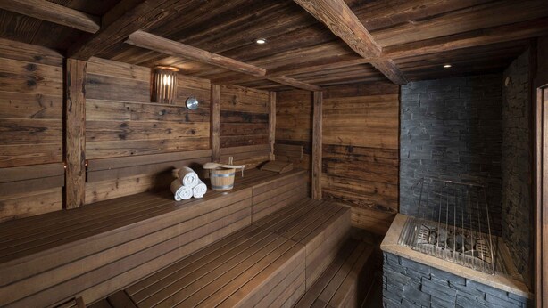 A wooden sauna with benches, a stone wall, and a metal grill. - Holiday apartment with wellness area (Engelberg)