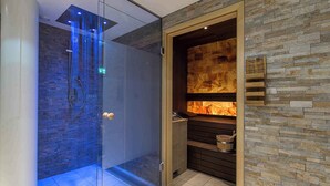 A modern bathroom with a glass-enclosed shower, a stone wall with a built-in fireplace, and a wooden shelf with a bucket. - Holiday apartment with wellness area (Engelberg)