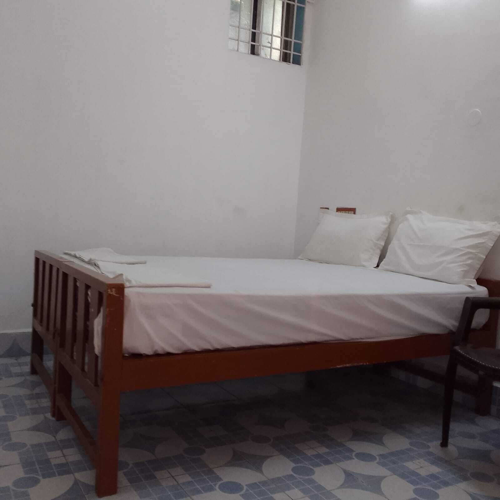 A single bed with a wooden frame and white bedding in a room with plain white walls and patterned floor tiles.