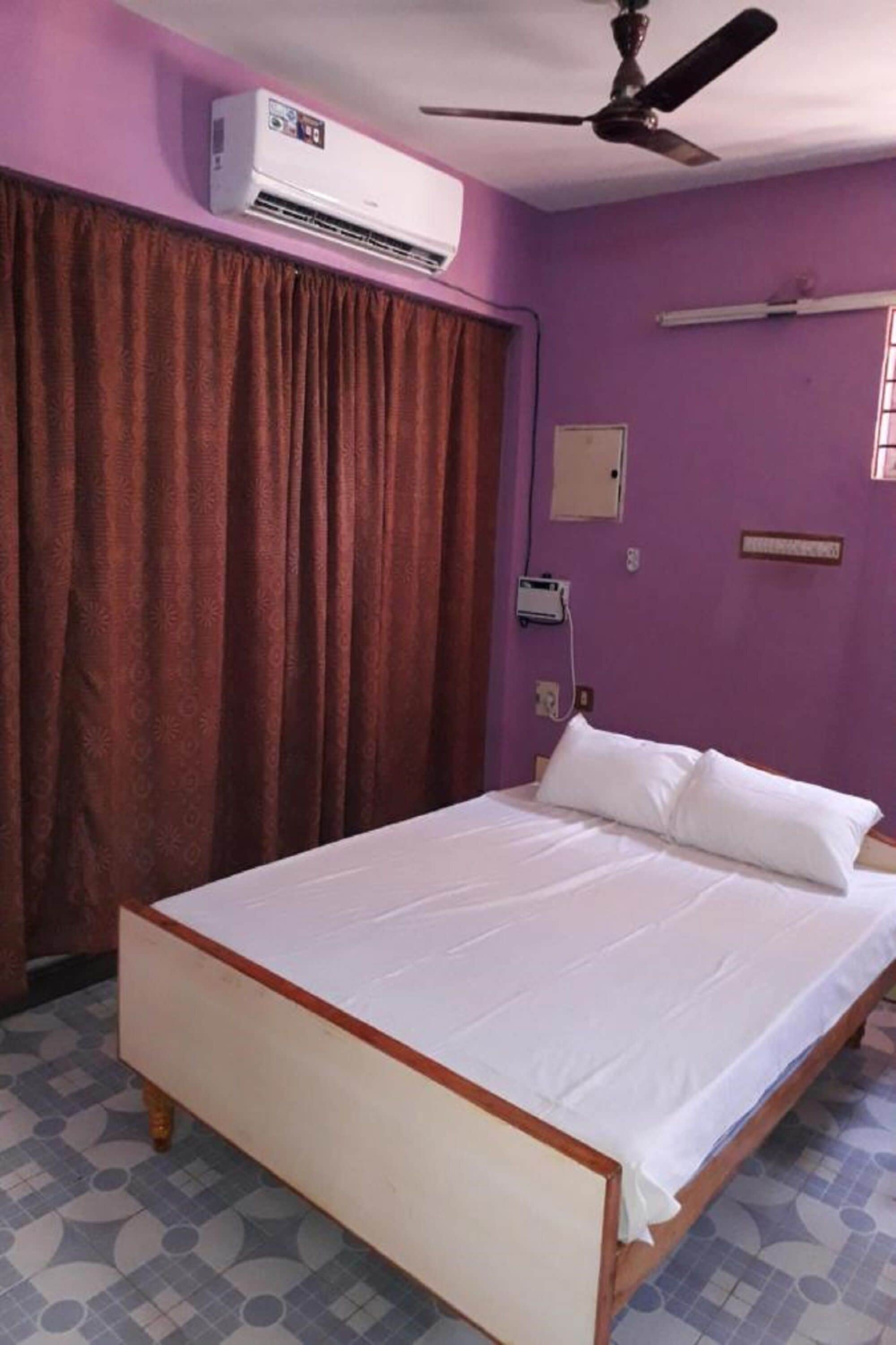 A room with a bed, an air conditioner, a ceiling fan, a window with curtains, and a wall-mounted electrical outlet.
