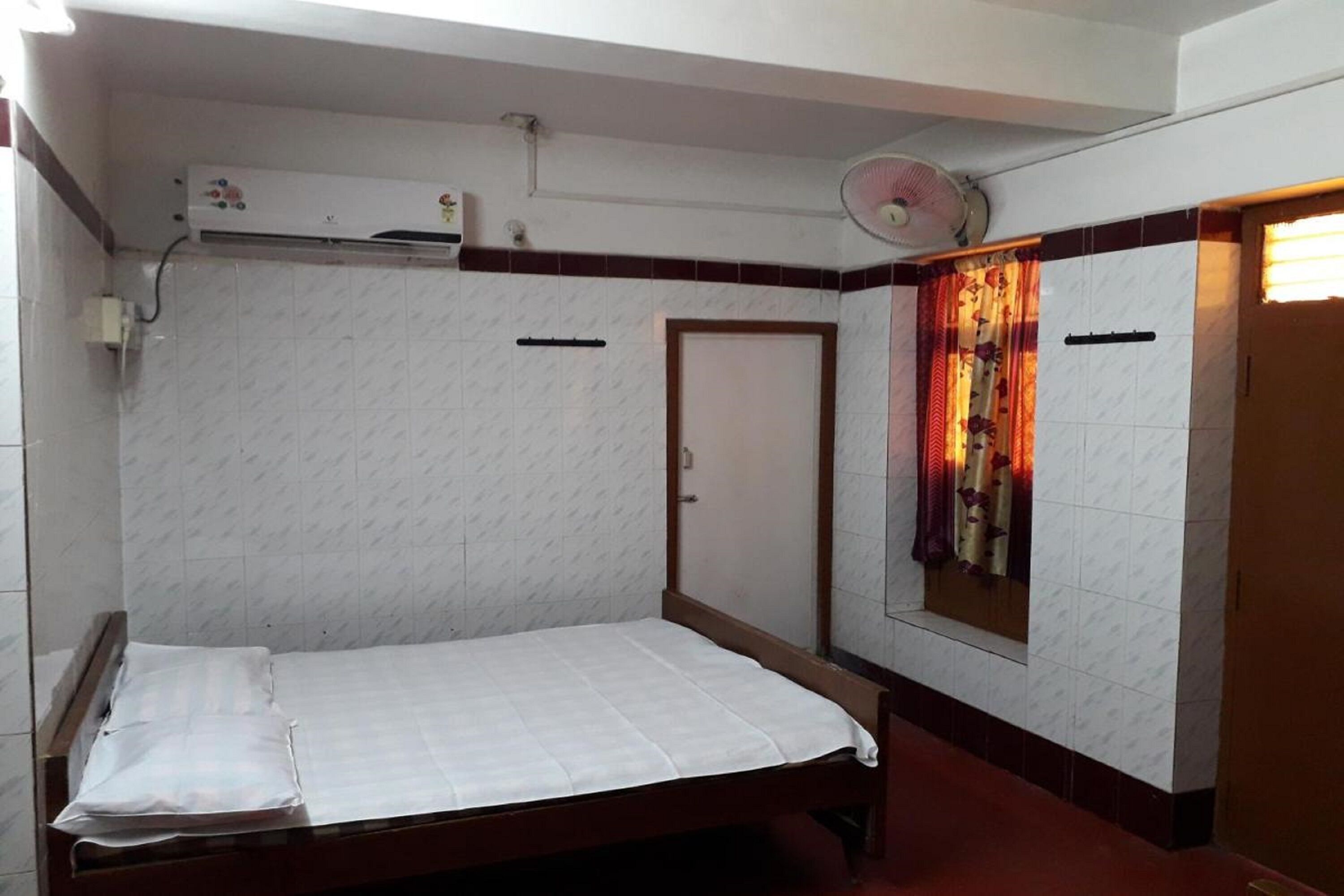 A single bed in a room with white patterned walls, a door, a window with curtains, and a ceiling fan.