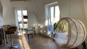 Une pièce spacieuse avec un plafond haut, un fauteuil suspendu, un coin repas avec des chaises et une table, et de grandes fenêtres laissant entrer la lumière naturelle.