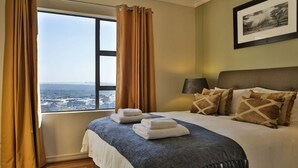 A hotel room with a large window, a bed with pillows, a lamp, and a framed picture on the wall. - Westay Westpoint Apartments (Johannesburg)