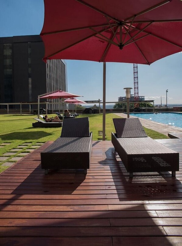 A wooden deck with two lounge chairs and a red umbrella, overlooking a swimming pool and a grassy area with a tall building in the background. - Westay Westpoint Apartments (Johannesburg)