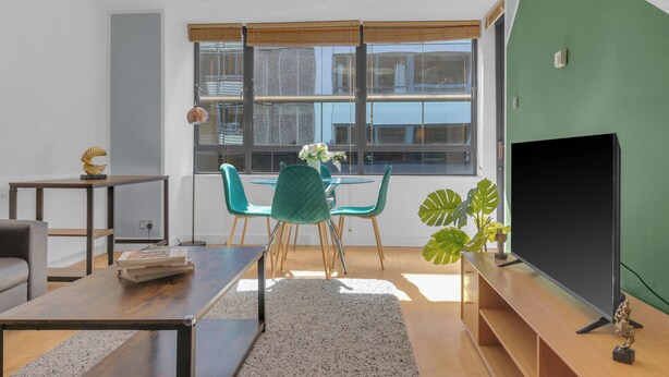 Una sala de estar moderna con una mesa de comedor, sillas, un sofá, un televisor y un amplio ventanal con persianas.