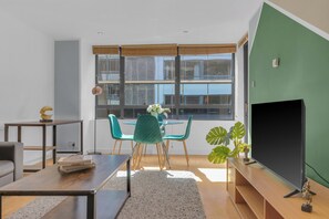 Una sala de estar moderna con una mesa de comedor, sillas, un sofá, un televisor y un amplio ventanal con persianas.