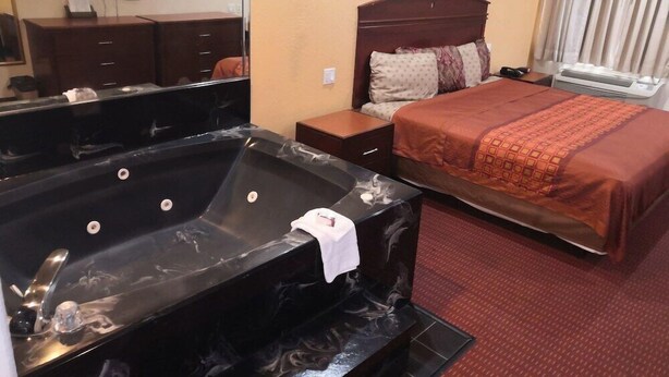A hotel room with a large black jacuzzi, a bed with a brown headboard, and a small wooden nightstand. - Scottish Inn & Suites IAH West (Houston)