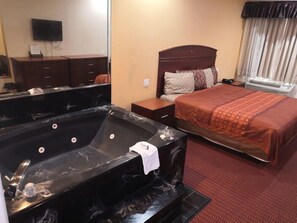 A hotel room with a large black jacuzzi, a bed with a brown headboard, and a small wooden nightstand. - Scottish Inn & Suites IAH West (Houston)