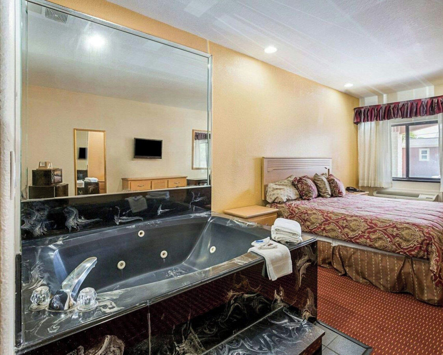 Habitación de hotel con una bañera grande, una cama con colcha estampada, un televisor y un espejo.