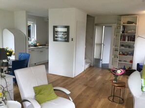 Ein modernes Wohnzimmer mit einem weißen Sofa, einem grünen Kissen und einem Holztisch. Ein Bücherregal voller Bücher und Dekorationsgegenstände, und eine Treppe, die in eine andere Ebene führt.