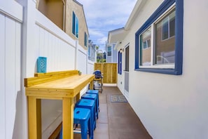 Une ruelle étroite avec une table en bois et des tabourets bleus, une clôture en bois et une fenêtre dotée d’un cadre bleu.