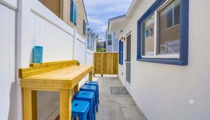 A narrow alleyway with a wooden table and blue stools, a wooden fence, and a small potted plant.