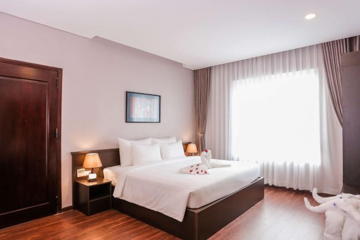 Habitación de hotel con una cama grande, dos mesitas de noche con lámparas, un armario de madera, un cuadro en la pared y un amplio ventanal con cortinas.