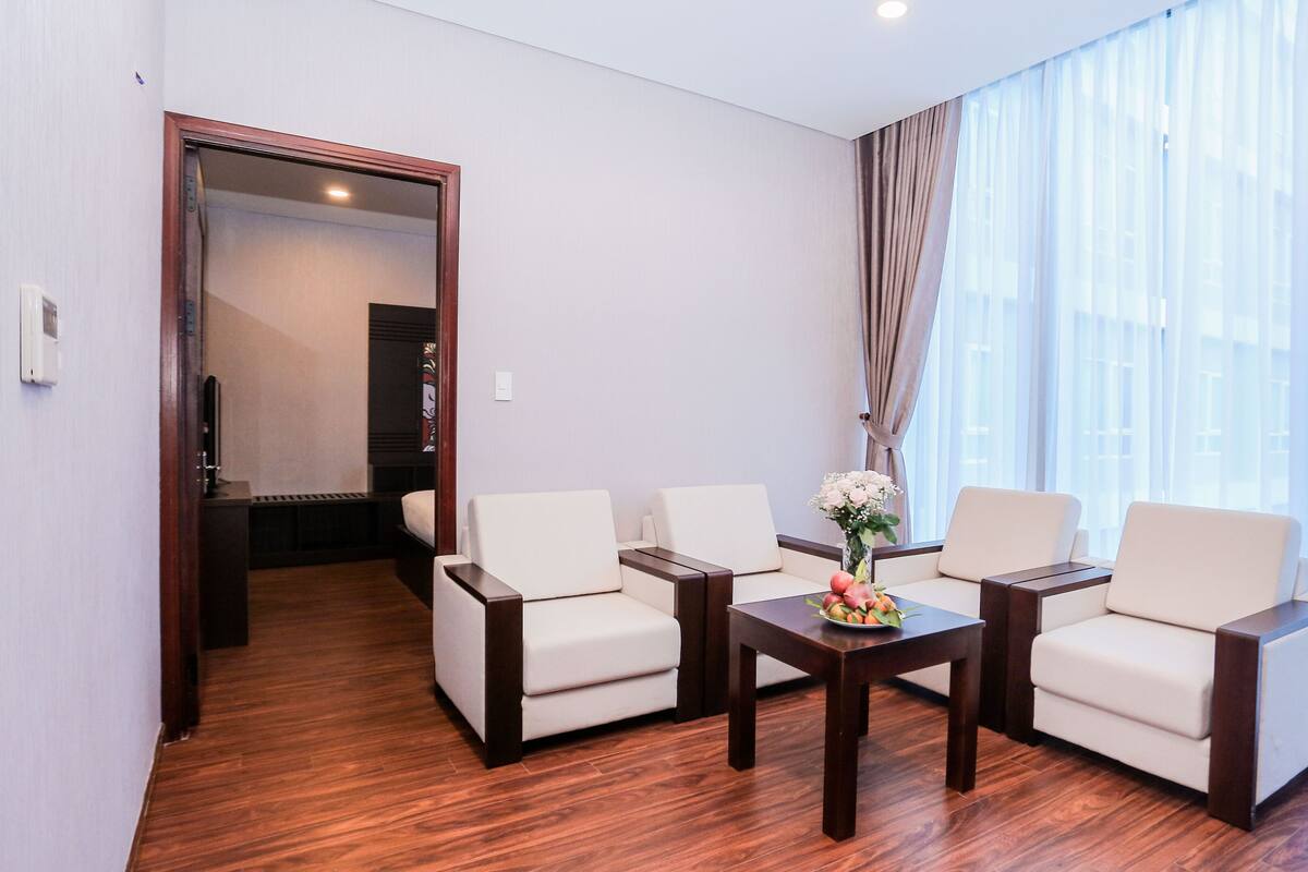 A modern hotel room with a wooden floor, a sofa set, a small table with a vase of flowers, and a large window with sheer curtains.
