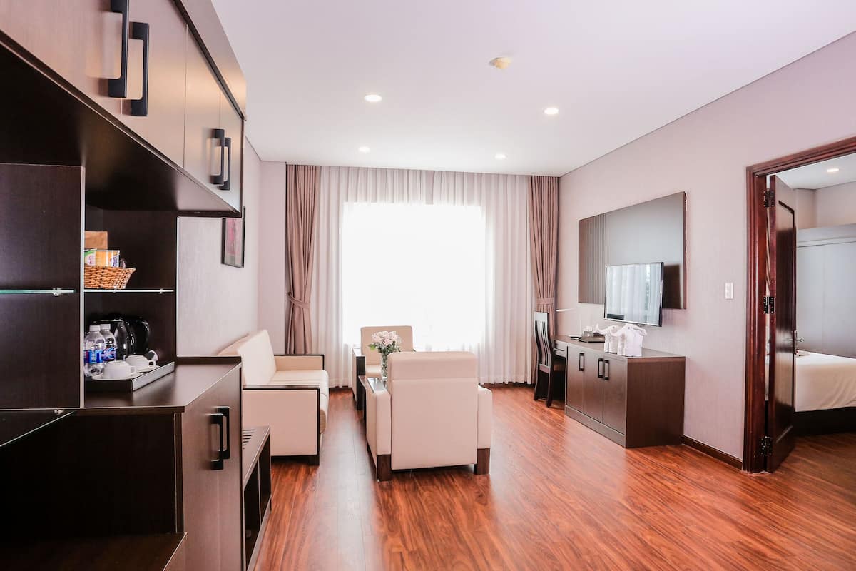 A hotel room with a wooden floor, a seating area with a sofa and chairs, a minibar, a large mirror, and a television.