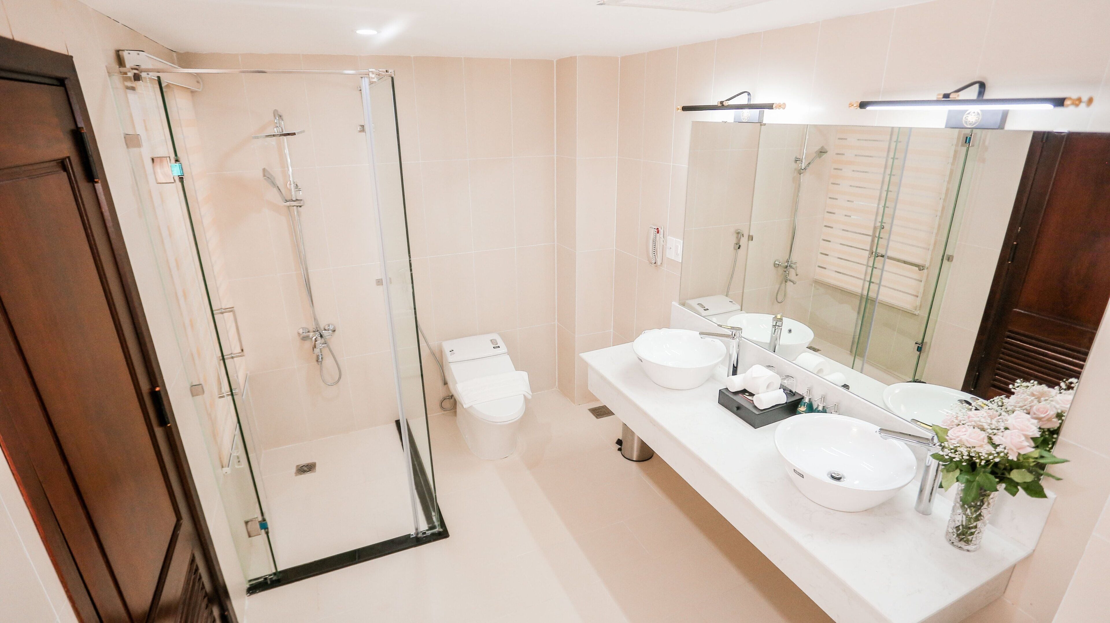 A modern bathroom with a glass-enclosed shower, two sinks, a large mirror, and a vase with flowers.