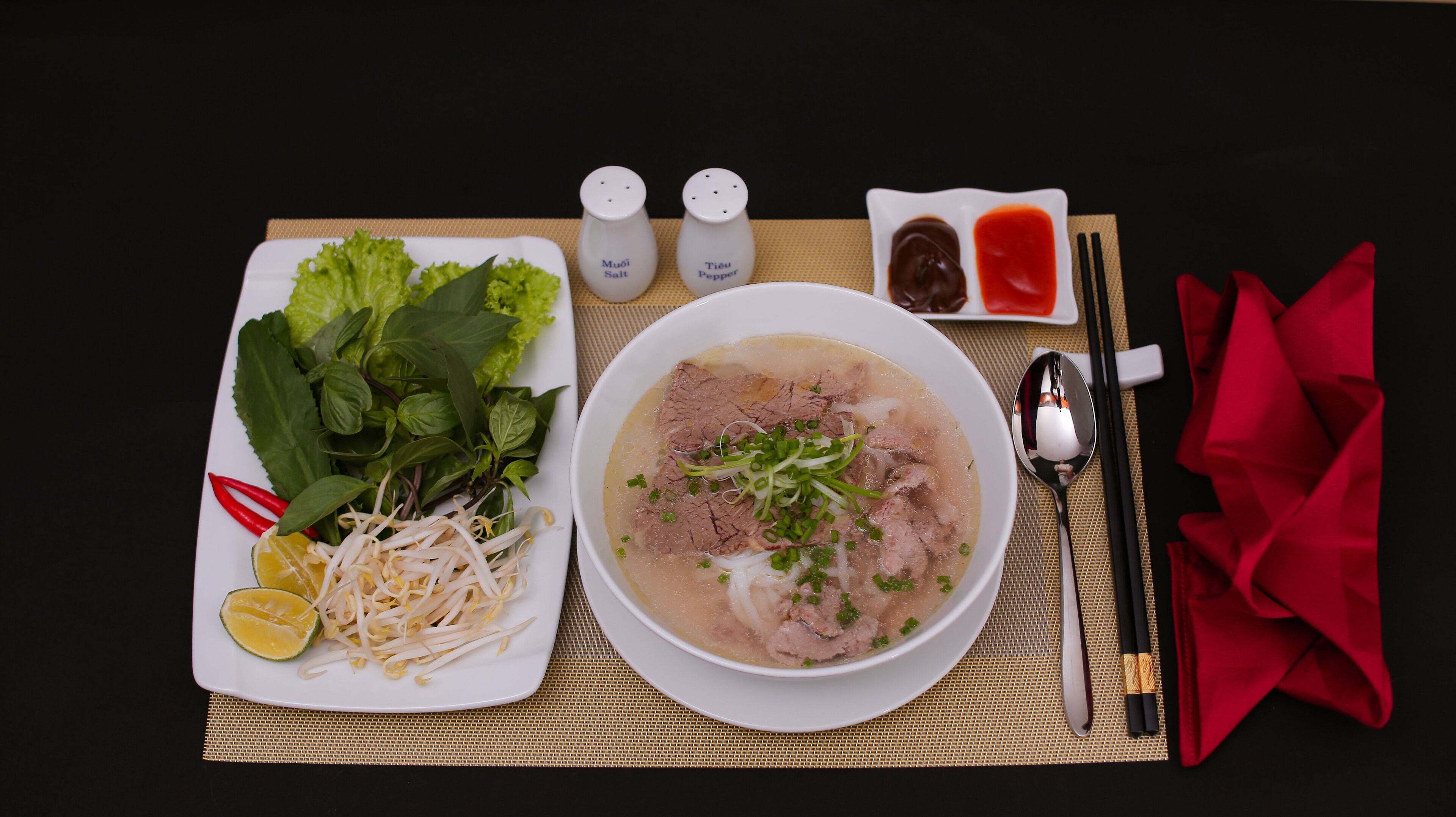 A bowl of soup with beef, vegetables, and herbs, accompanied by a plate of fresh herbs, bean sprouts, and lime.