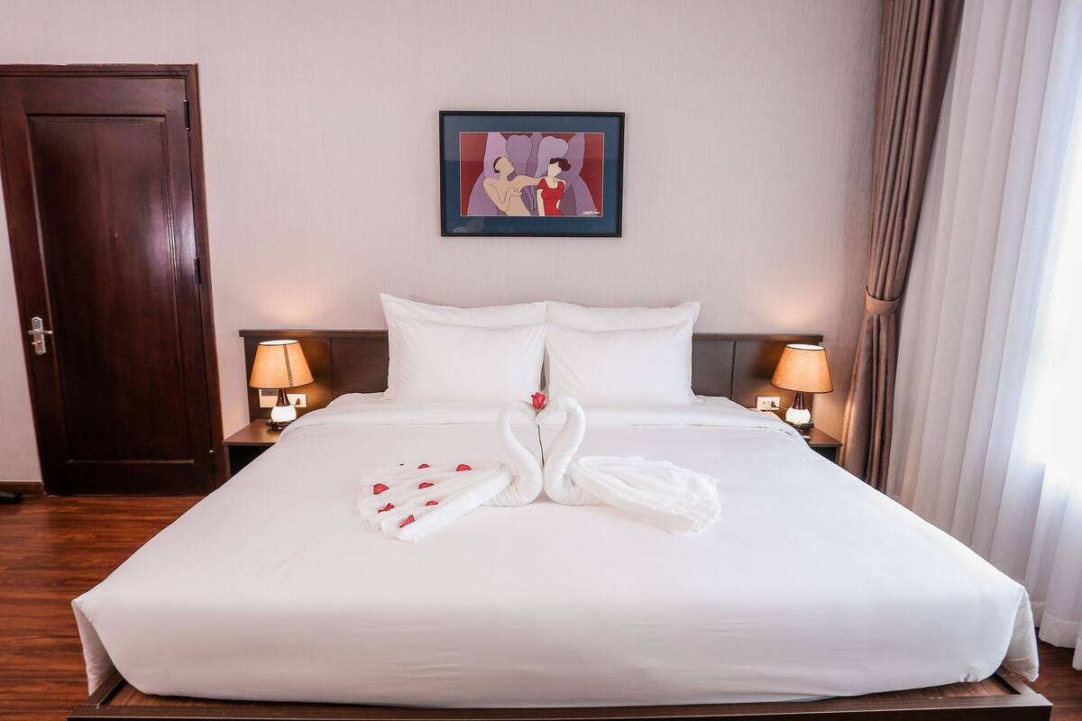 A hotel room with a large bed, white linens, two bedside lamps, a framed artwork on the wall, and a wooden door.