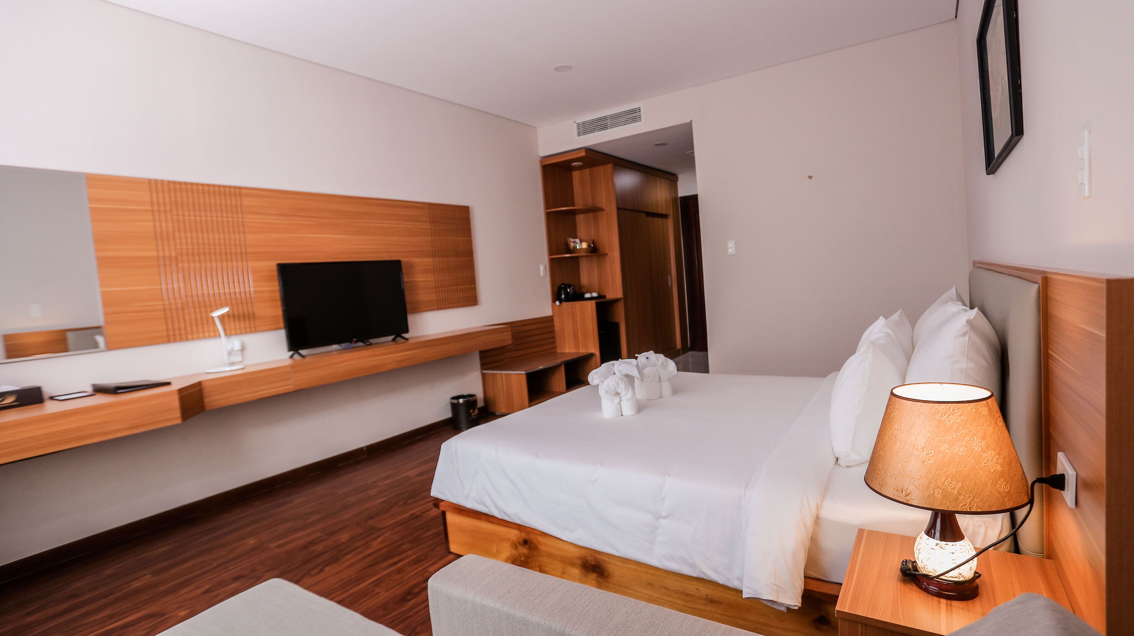 A hotel room with a bed, a TV on a wooden shelf, a bedside table with a lamp, and a wardrobe.