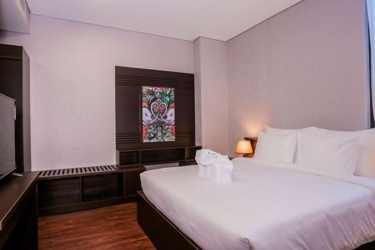 Una habitación de hotel con una cama grande, televisión, un ventanal decorativo con vitrales y una mesita de noche con lámpara.