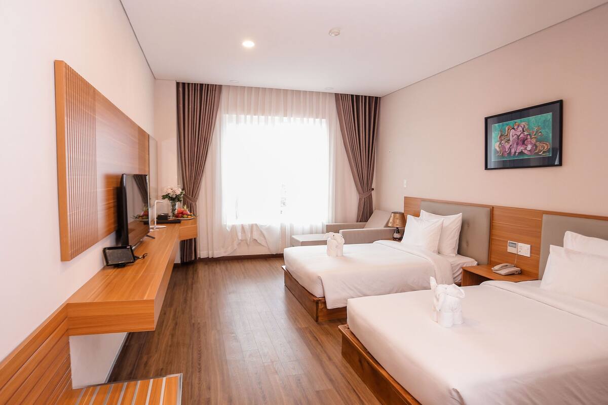 A hotel room with two beds, a desk with a flat-screen TV, a bedside table with a lamp, and a painting on the wall.