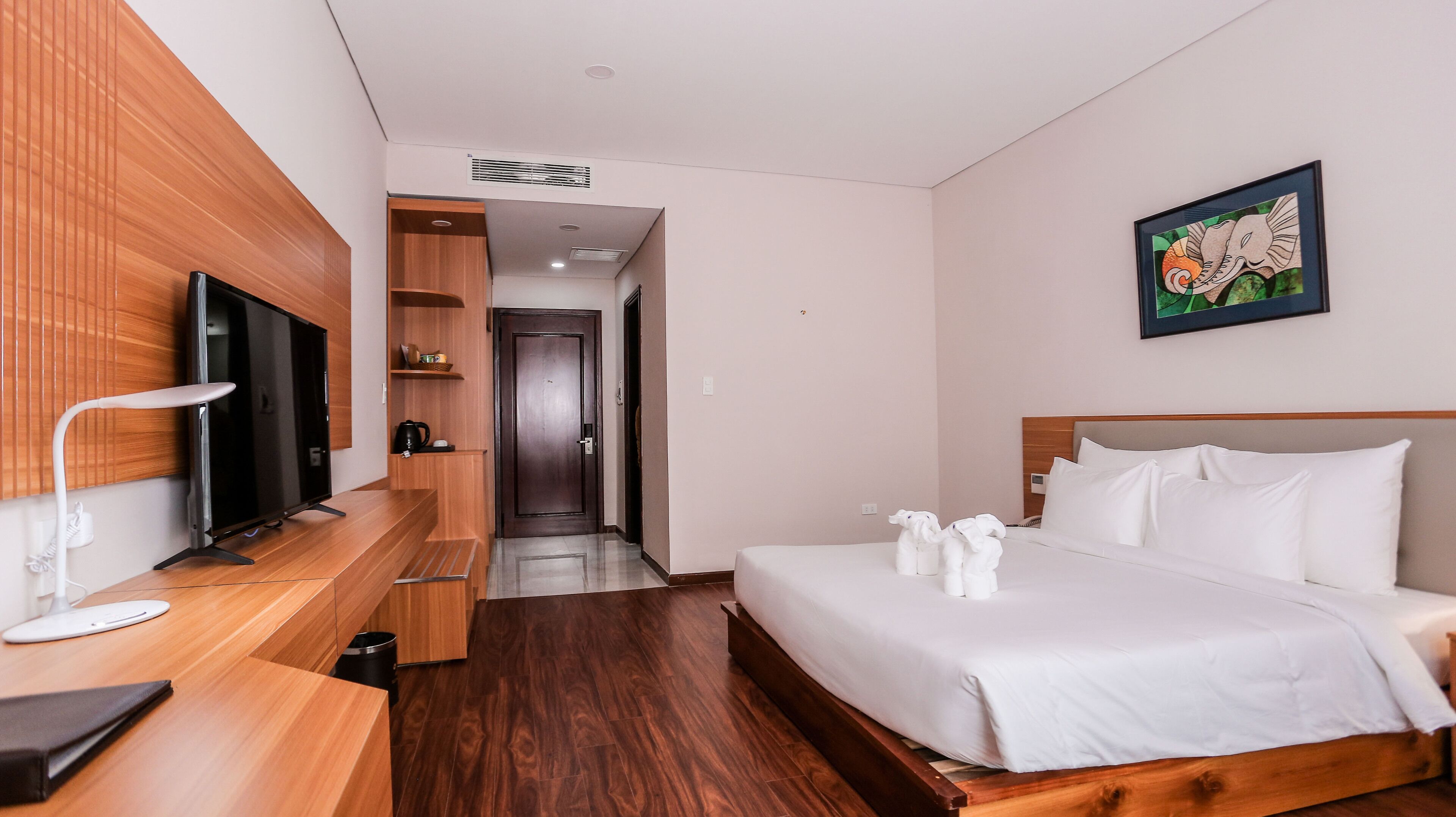 Une chambre d’hôtel avec un bureau en bois, une télévision, un lit avec du linge de maison blanc et un tableau au mur.