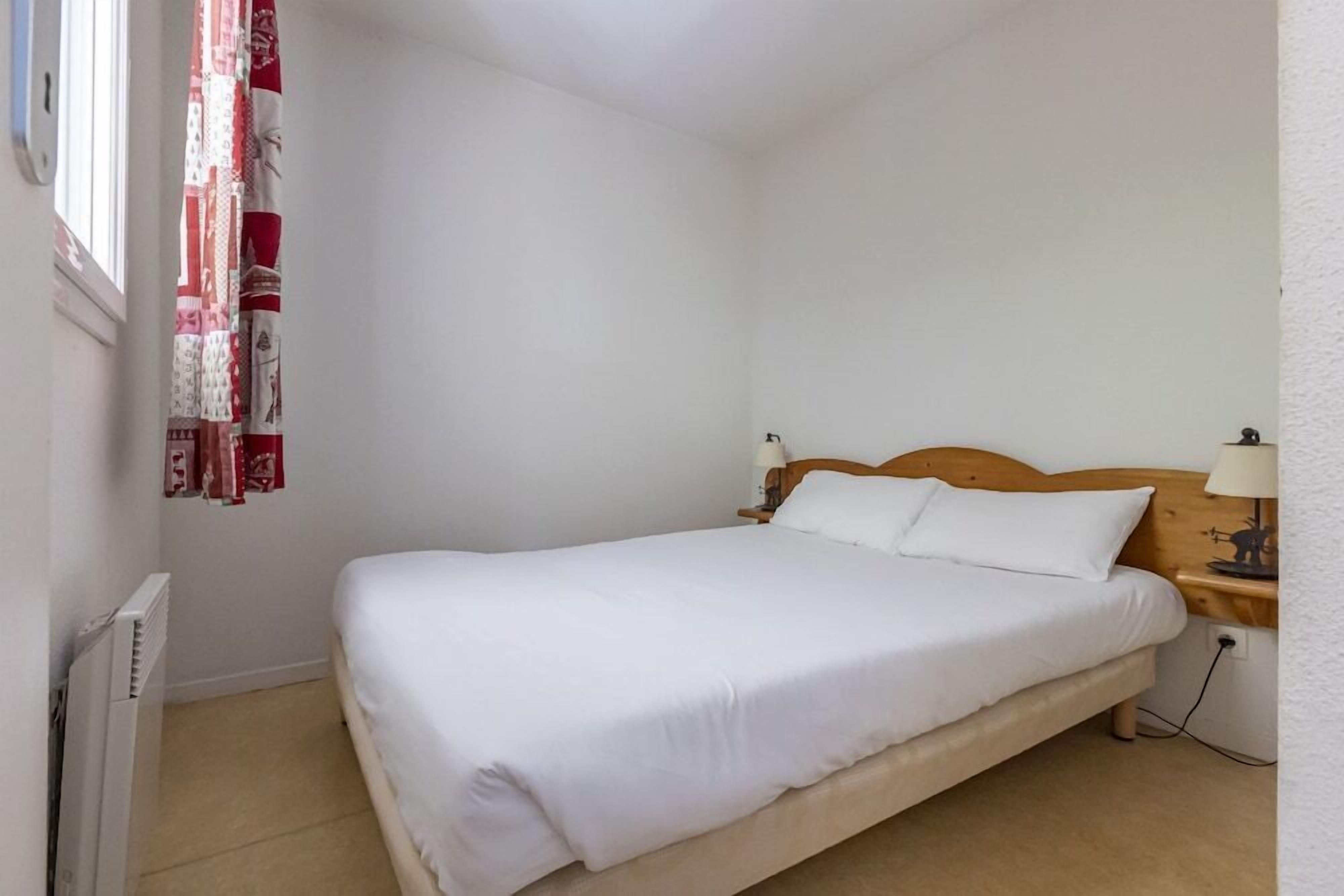 Una cama individual con ropa de cama blanca en una habitación con paredes blancas, un cabecero de madera, una mesita de noche con lámpara y una ventana con cortinas rojas y blancas.