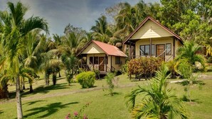 Un hôtel de style bungalow, avec un toit rouge et des murs de couleur beige, entouré d’une végétation luxuriante et de palmiers.