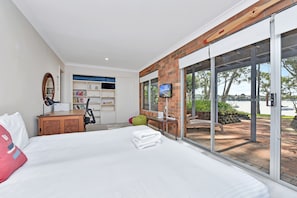 A modern bedroom with a large bed, a desk, a television, and a view of a body of water through large sliding doors.