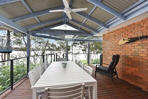 A covered outdoor seating area with a table and chairs, a ceiling fan, and a view of trees and a brick wall.