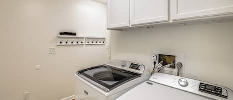 A compact laundry room with a front-loading washer and dryer, a white wall with a shelf above the dryer, and a wooden floor.