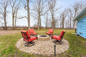 A circular seating area with four red chairs around a stone fire pit, set on a gravel surface with trees in the background.