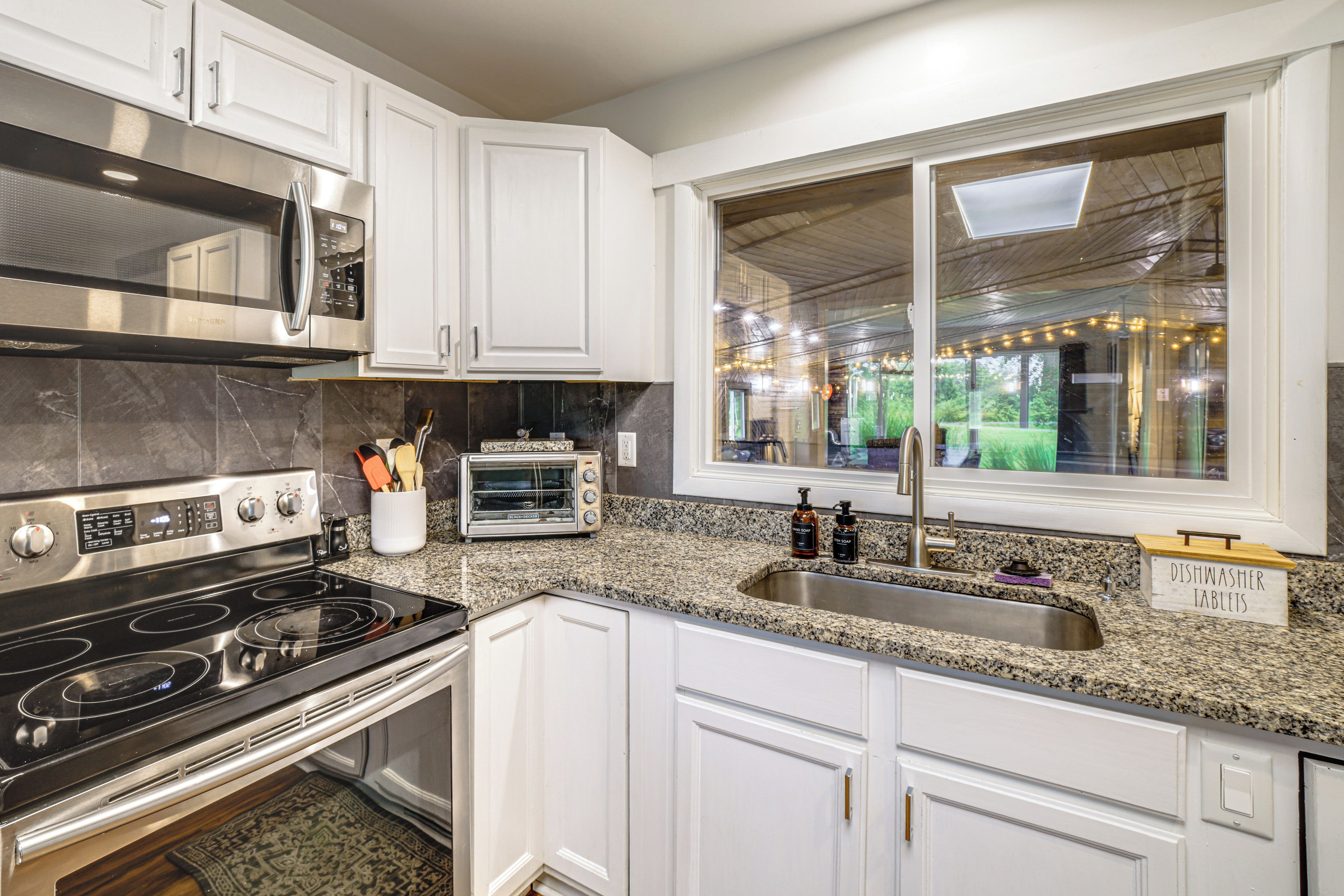 A modern kitchen with white cabinetry, a stainless steel stove, a microwave, and a window with a view of greenery.