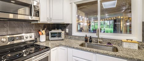 A modern kitchen with white cabinetry, a stainless steel stove, a microwave, and a window with a view of greenery.