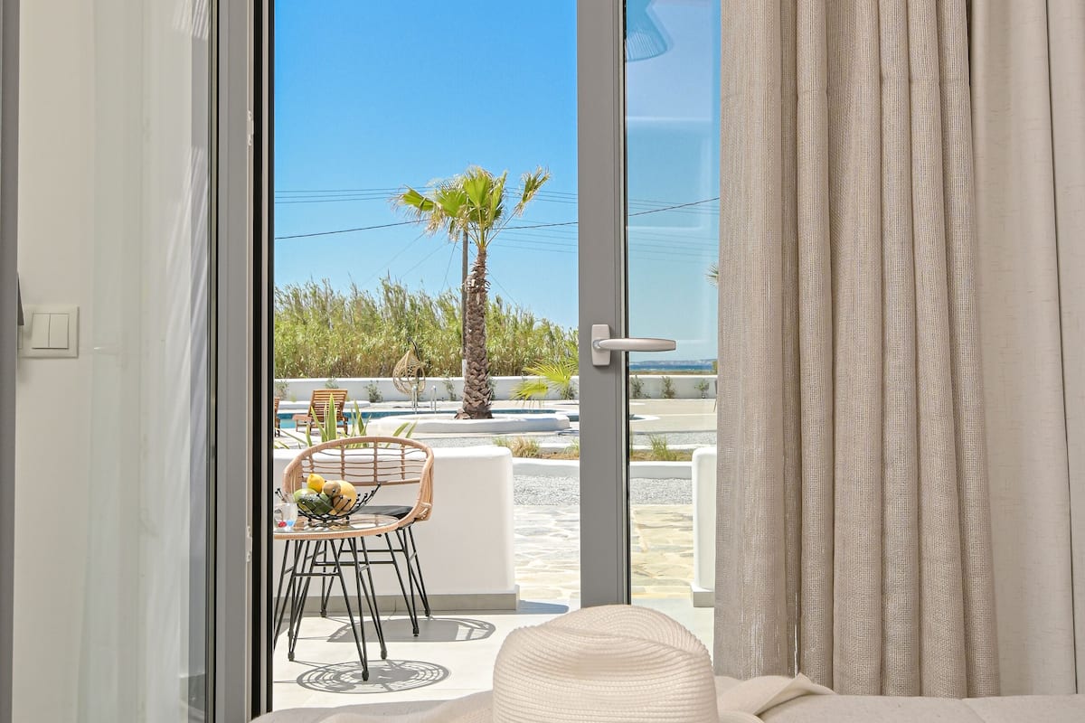 Un dormitorio con puerta de cristal corredera que da a un balcón con mobiliario exterior y una palmera.