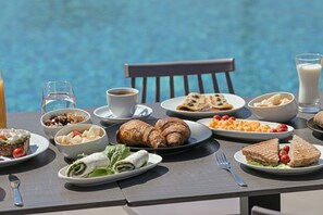 A table set with breakfast items including croissants, sandwiches, and fruit, overlooking a pool. - Naxos Finest Hotel & Villas (Naxos)