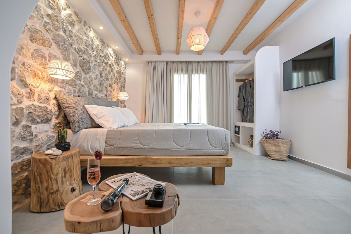 Un dormitorio con pared de piedra, vigas de madera, cama, televisor y una pequeña mesa con una copa de vino.
