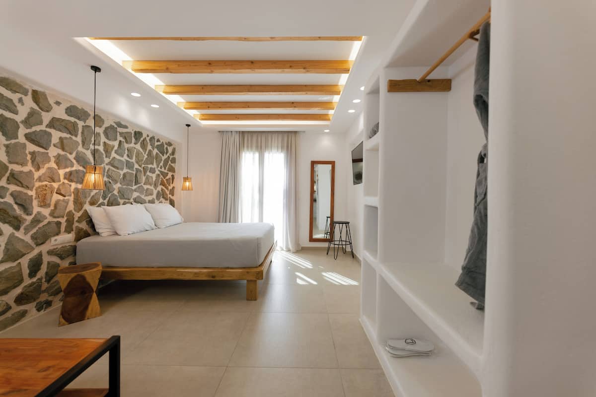 Un dormitorio moderno con pared de piedra, vigas de madera en el techo, una cama, una mesita de noche y un armario suspendido.