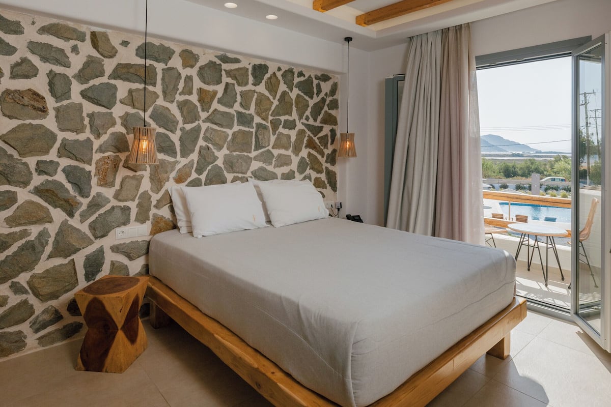 Un dormitorio con pared de piedra, vigas de madera, una cama, una lámpara colgante y vistas a través de un ventanal.