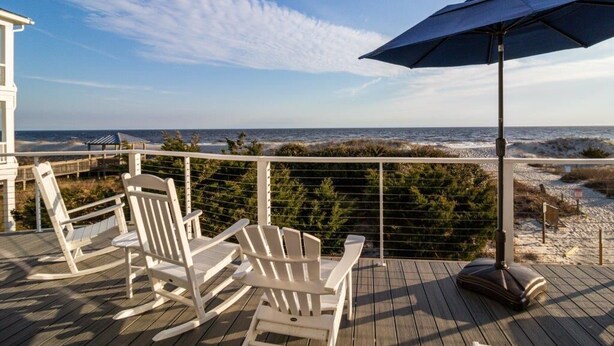 Une terrasse en bois avec des fauteuils Adirondack blancs et un parasol bleu, donnant sur une plage et l’océan.