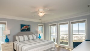 Une chambre à coucher avec un lit, deux tables de chevet avec des lampes, un ventilateur de plafond et de grandes fenêtres donnant sur la plage.