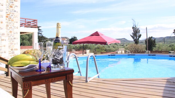 Eine Holzterrasse mit einem Tisch, auf dem eine Champagnerflasche, Gläser und eine Bananenschale stehen. Im Hintergrund ein Swimmingpool mit einem roten Sonnenschirm und Blick auf Hügel.