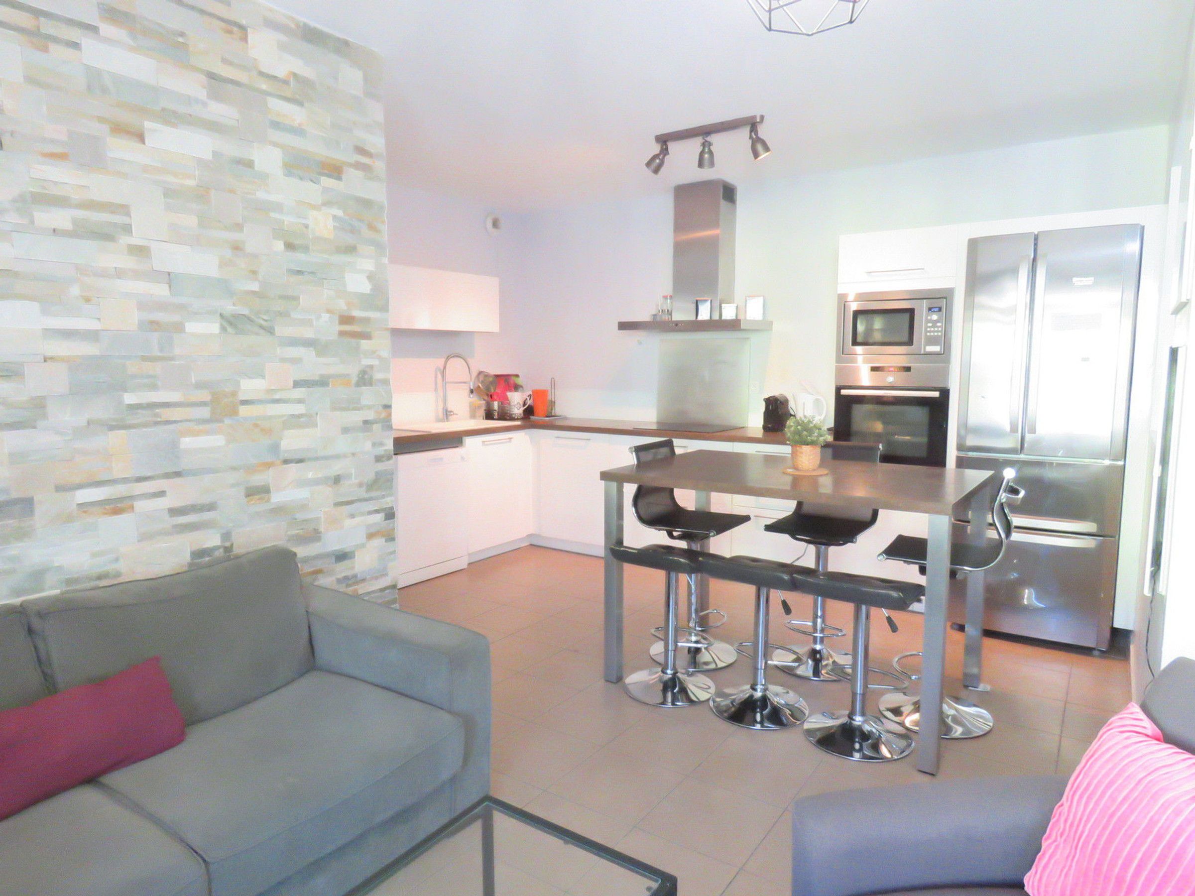 A modern kitchen with stainless steel appliances, a dining area with bar stools, and a grey sofa with a pink pillow.